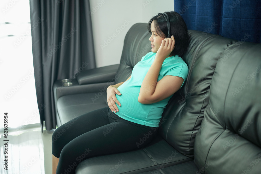Asian pregnant woman listening to music and sitting on sofa in room