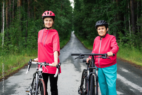 Mother, daughter with bicycles in sportive helmets, suits look at camera, standing on road surrounded by trees. Young sports coach and senior woman are going to ride bicycles in forest