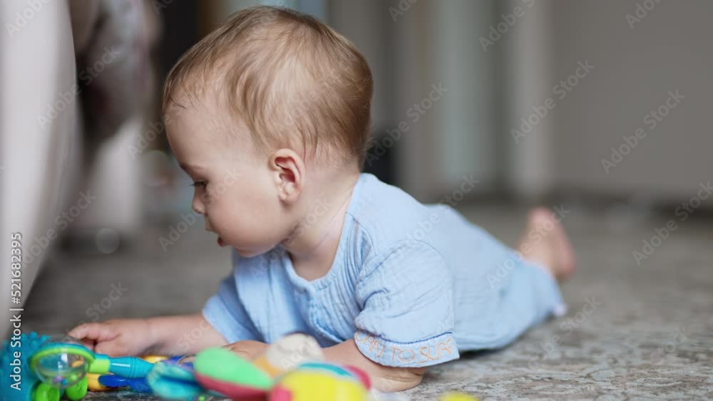 Nice kid wearing blue shirt lies on the floor. Little boy touches toys lying beside him. Close up. Blurred backdrop. Nice kid wearing blue shirt lies on the floor. Little boy touches toys lying beside him. Close up. Blurred backdrop.
