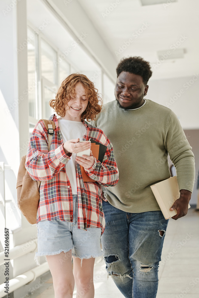 Two teenage classmates in casualwear discussing curious stuff in smartphone held by cute smiling girl after lessons Two teenage classmates in casualwear discussing curious stuff in smartphone held by cute smiling girl after lessons
