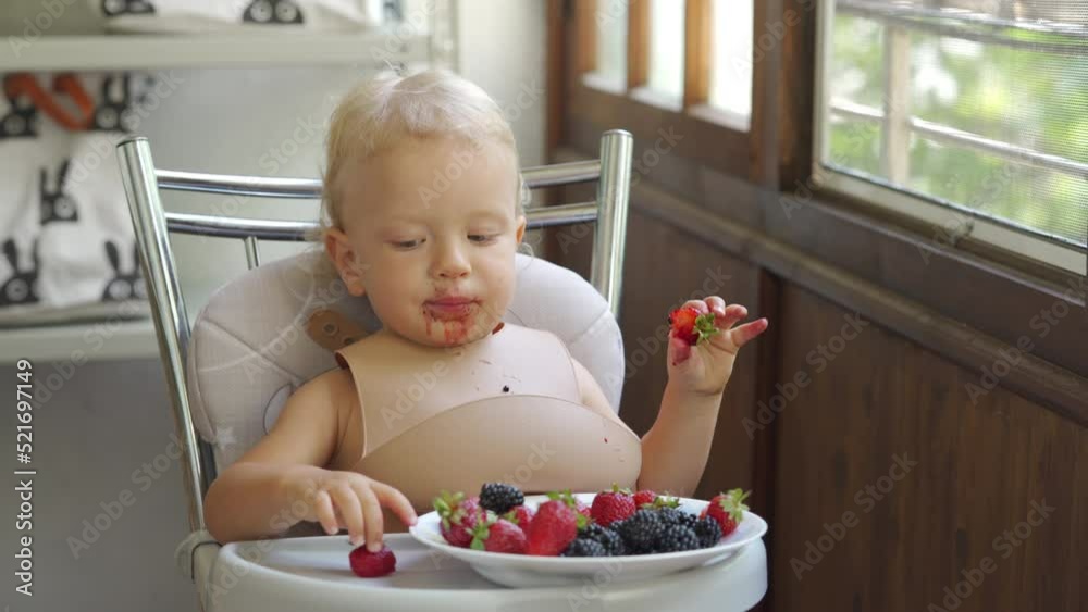 Baby boy eating berries for the first time, 1.5 year old toddler child sitting on highchair eatsstrawberries blackberries blueberries on the balcony in summer. Baby boy eating berries for the first time, 1.5 year old toddler child sitting on highchair eatsstrawberries blackberries blueberries on the balcony in summer.