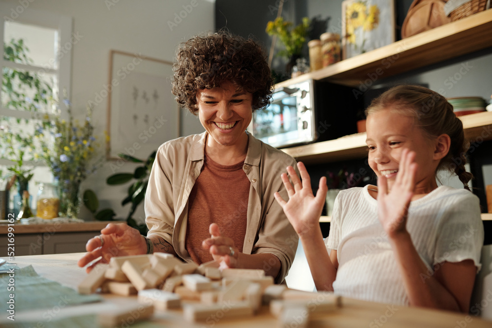 Young mother and cute daughter laughing at destroyed tower from wooden blocks on table while playing leisure game at home Young mother and cute daughter laughing at destroyed tower from wooden blocks on table while playing leisure game at home