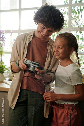 Young smiling woman in casualwear showing new photos taken on photocamera to her cute youthful daughter at leisure