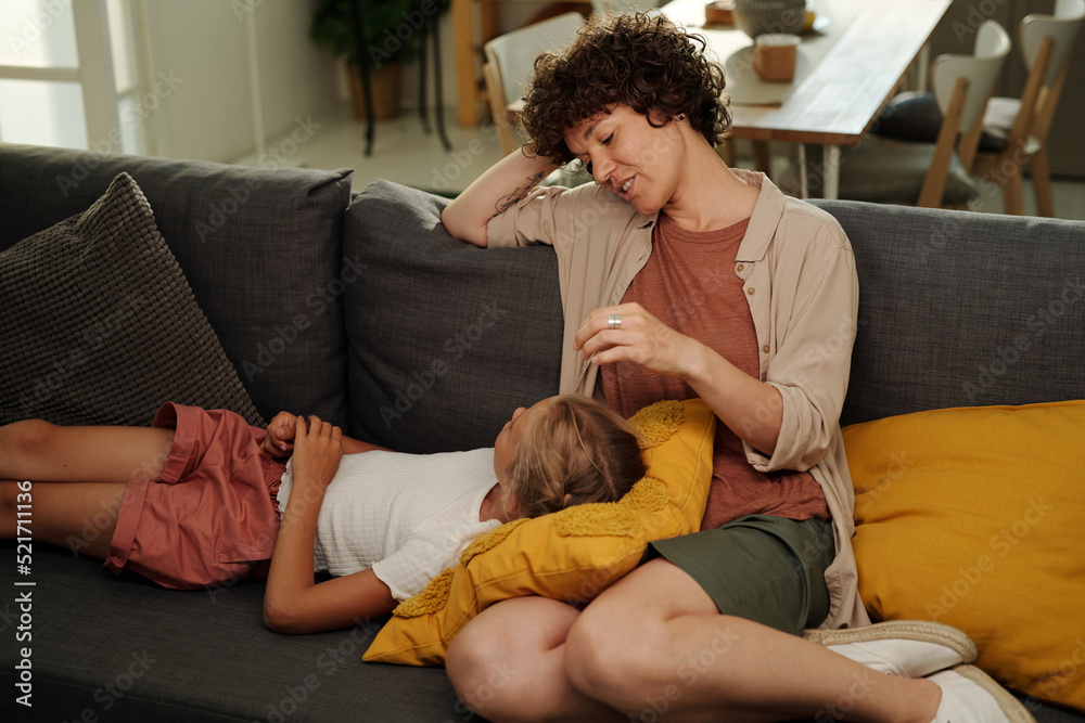 Youthful serene girl lying on pillow on her mother knees and both having chat while relaxing on comfortable couch in living room Youthful serene girl lying on pillow on her mother knees and both having chat while relaxing on comfortable couch in living room
