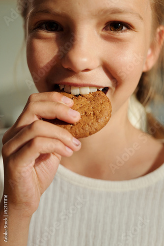 Happy youthful girl biting fresh tasty homemade cookie of round shape while standing in front of camera and looking at you