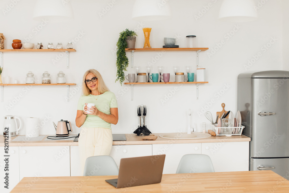 Modern attractive middle-aged woman drinking coffee and working on laptop in kitchen at home Modern attractive middle-aged woman drinking coffee and working on laptop in kitchen at home