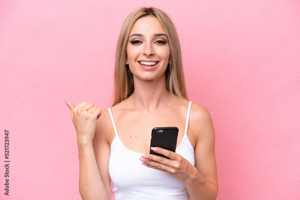 Pretty blonde woman isolated on pink background using mobile phone and pointing to the lateral Pretty blonde woman isolated on pink background using mobile phone and pointing to the lateral