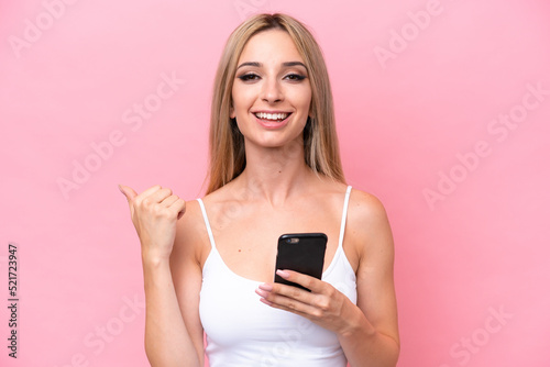 Pretty blonde woman isolated on pink background using mobile phone and pointing to the lateral