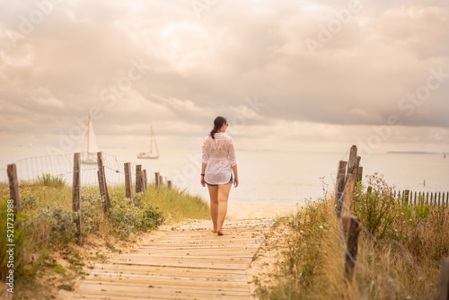 A young woman walks on the wooden pontoon that leads to the beach. In the background you can see two boats with white sails, sailing in the calm water of the ocean. The fence forms a path to the beach