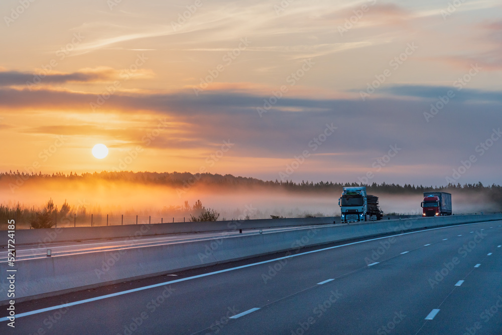 Landscape of a highway at dawn with the sun beginning to rise, two trucks circulating and the mist tucked between the trees.