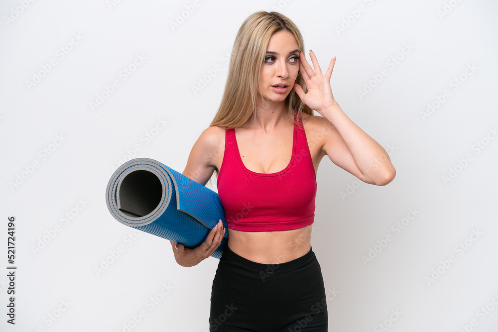 Young sport blonde woman going to yoga classes while holding a mat isolated on white background listening to something by putting hand on the ear