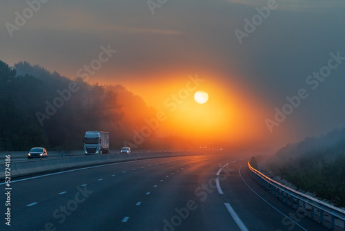 Landscape of a highway with a truck and several vehicles circulating at dawn, and the sun appearing through the clouds, allowing its circumference to be seen through the mist.