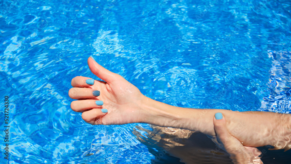 Close-up of female manicured hands. Woman's hands with bright glitter blue fingernails inside of bright blue swimming pool water.