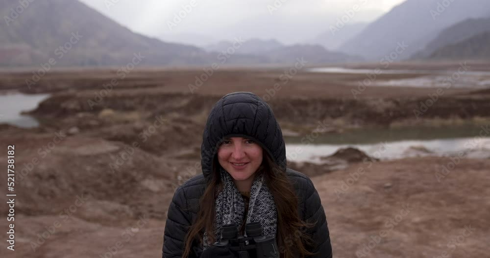 A woman is walking through the mountains with her binoculars and looks at the camera smiling. Concept of tourist, happiness, tourism, natural environment, environmental awareness A woman is walking through the mountains with her binoculars and looks at the camera smiling. Concept of tourist, happiness, tourism, natural environment, environmental awareness