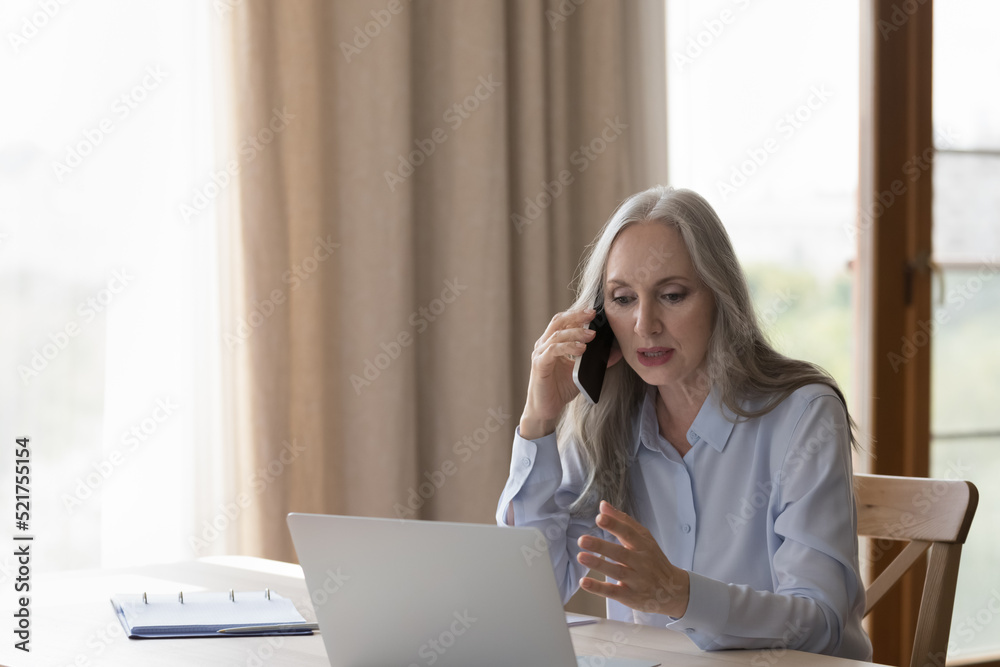 Focused busy older mature business leader woman talking to client on telephone, using online app on laptop for conversation, looking at display, sitting at home office table, speaking on smartphone Focused busy older mature business leader woman talking to client on telephone, using online app on laptop for conversation, looking at display, sitting at home office table, speaking on smartphone