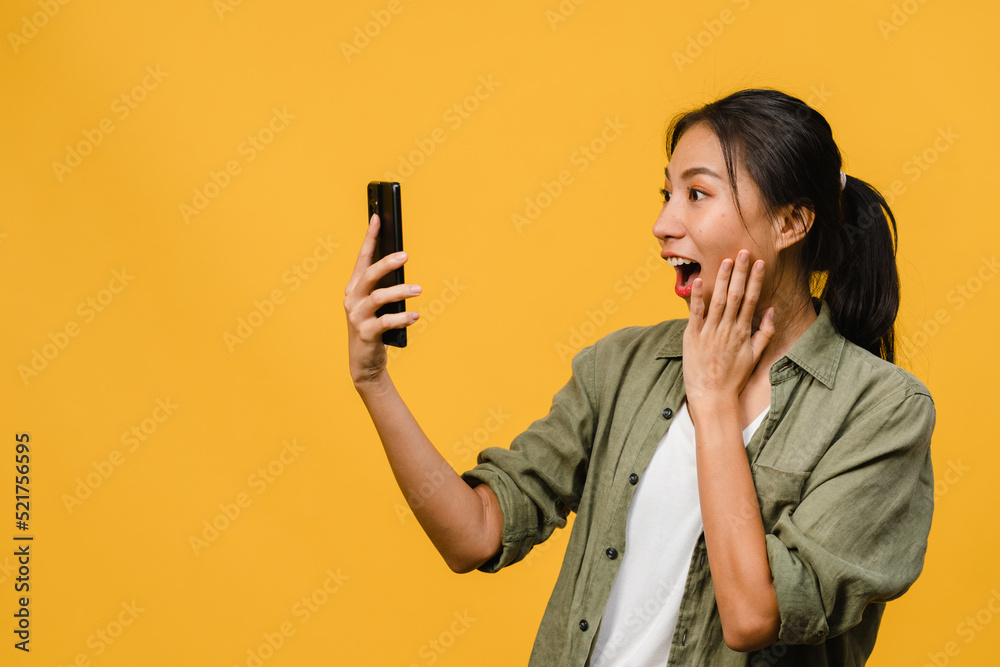 Surprised young Asia lady using mobile phone with positive expression, smiles broadly, dressed in casual clothing and stand isolated on yellow background. Happy adorable glad woman rejoices success. Surprised young Asia lady using mobile phone with positive expression, smiles broadly, dressed in casual clothing and stand isolated on yellow background. Happy adorable glad woman rejoices success.