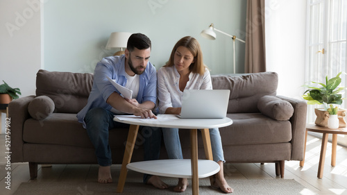 Serious millennial couple sit on sofa sorting out papers, reviewing bills for utility expenses, analysing taxes, do household payment use e-bank application on laptop. Manage finances concept