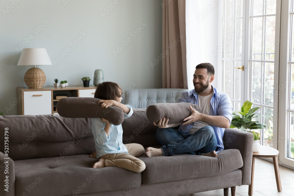 Little boy hold cushion play fight with young cheery father sit on sofa at home, happy family enjoy active pastime, spend leisure together at modern fashionable home. Games, fun, fatherhood concept