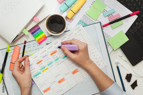Student desk. The girl makes notes on stickers in the study schedule. The process of homeschooling or preparing homework . Top view of the table with student stationery and notes.