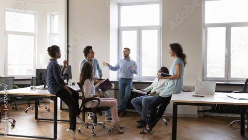 Multiethnic staff members gathered in coworking solve business, listen to male team leader during morning briefing in modern company office. Boss share information, corporate goals, tell about project