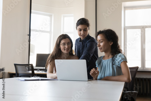 Three diverse young female colleagues, discuss new project, develop business strategy using laptop, prepare on-line presentation, planning collaborative task looking motivated. Teamwork, tech concept