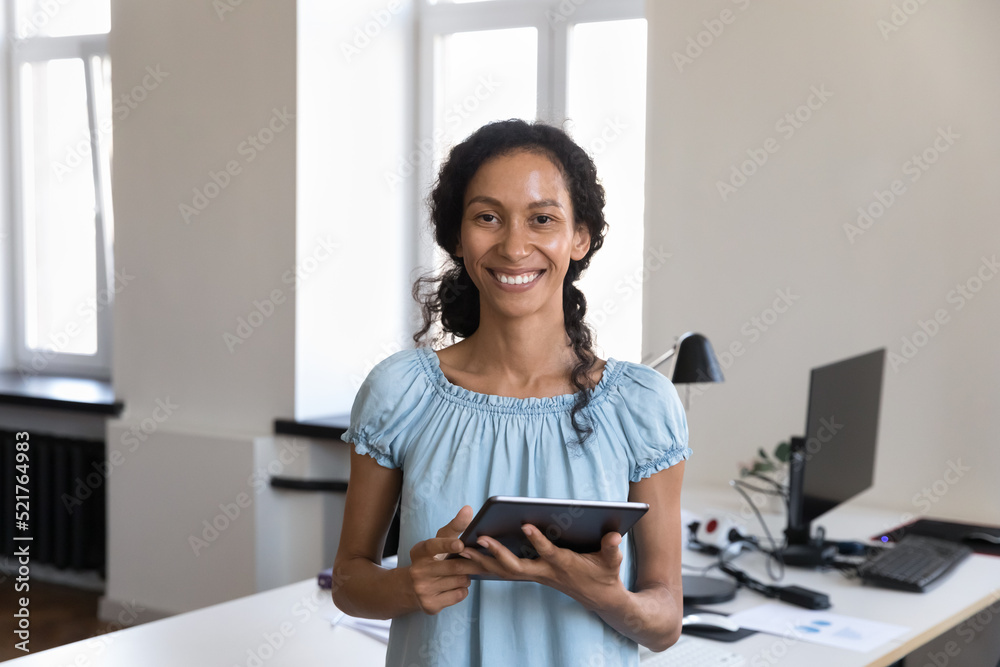 Obraz premium Young 35s African businesslady pose at workplace with digital tablet in hands smile look at camera. Workflow use modern wireless tech, apps, remote communication to client, corporate messaging concept
