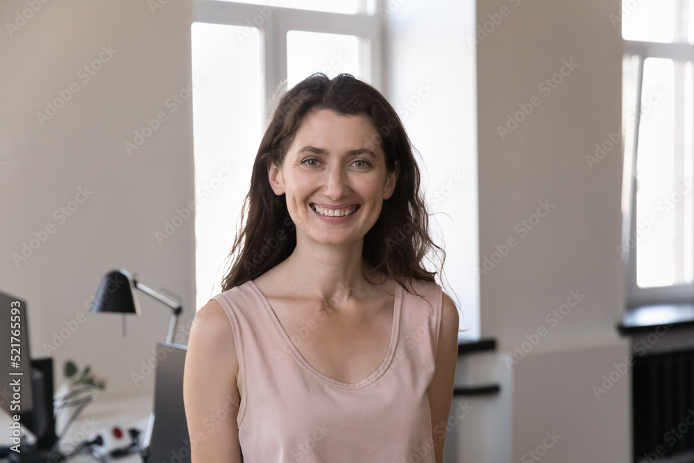 Attractive 35s businesswoman pose at modern workplace, head shot. Company female employee standing alone in office workspace staring at camera, career, workforce, professional worker portrait concept