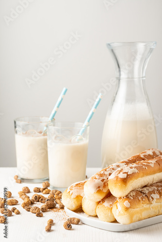 Close-up of bottle and glasses of horchata with straw, on table with tiger nuts and fartons, white background, vertical photo