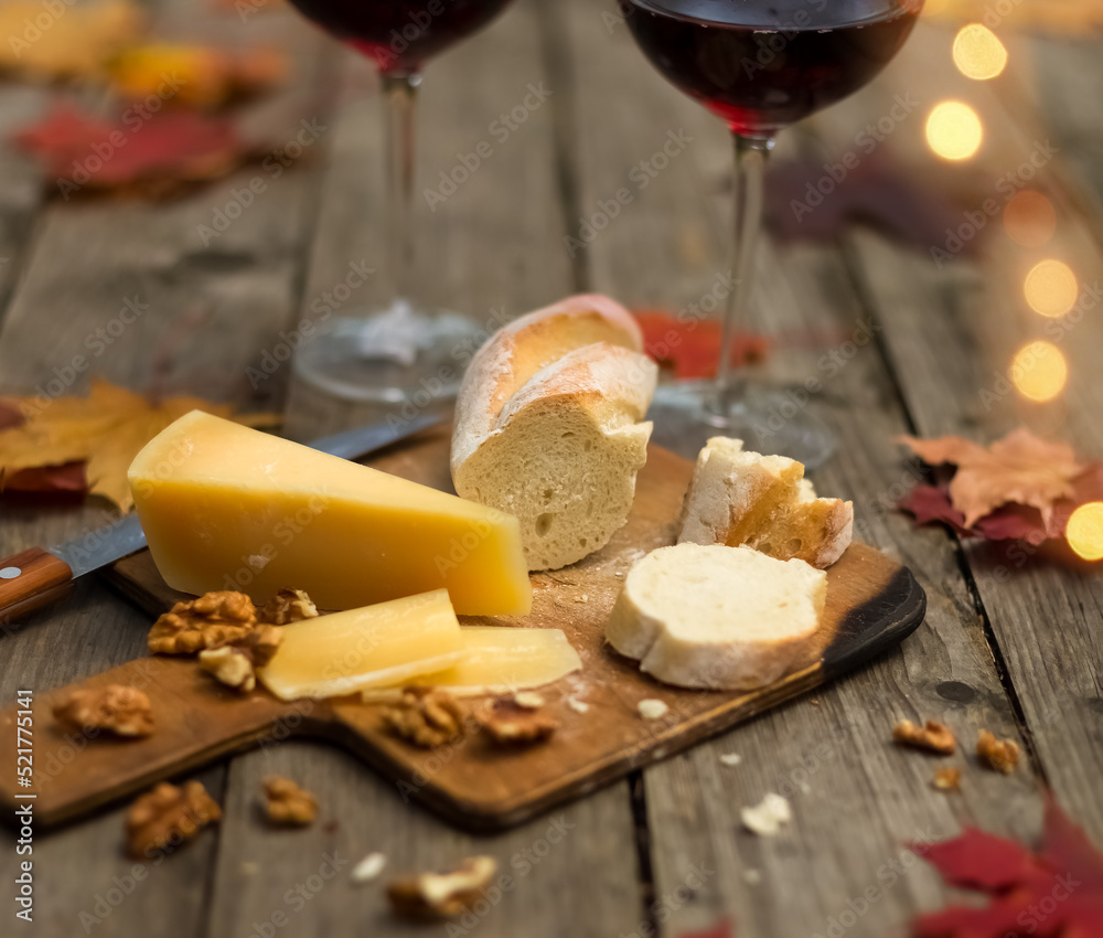 Close up of glasses of red wine with bread and cheese on wooden table with maple leaves. Autumn still life in evening outdoors