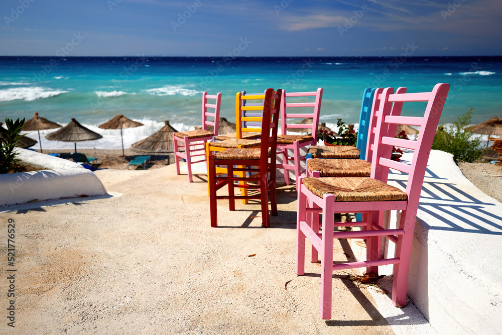 Multicoloured chairs against the backdrop of the green-blue sea and the dramatic sky of the Albanian Riviera. Postcard from the Mediterranean. Outdoor, Pink, yellow, brown chairs on white floor.