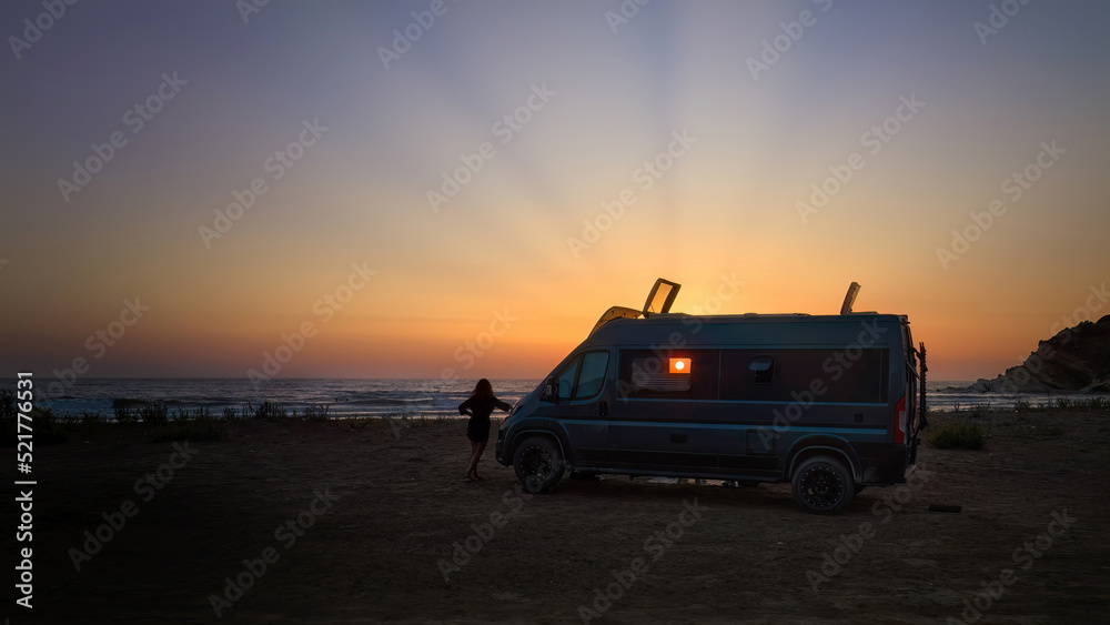 Silhouette of a offroad caravan and a young woman against the setting sun, the beach and the sea. Adventure of wild camping, nomadic life. Travelling in a van camper.