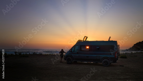 Silhouette of a offroad caravan and a young woman against the setting sun, the beach and the sea. Adventure of wild camping, nomadic life. Travelling in a van camper.