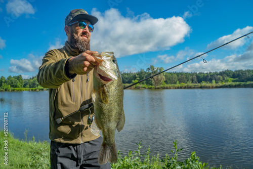 Bass fishing. Large bass fish in hand of pleased bearded fisherman with tackle. Largemouth perch at pond