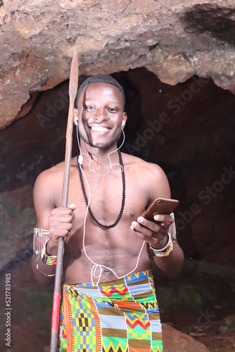A cave in Africa: Masai Moran warrior with a spear smiling listening to music on a mobile phone using earphones 