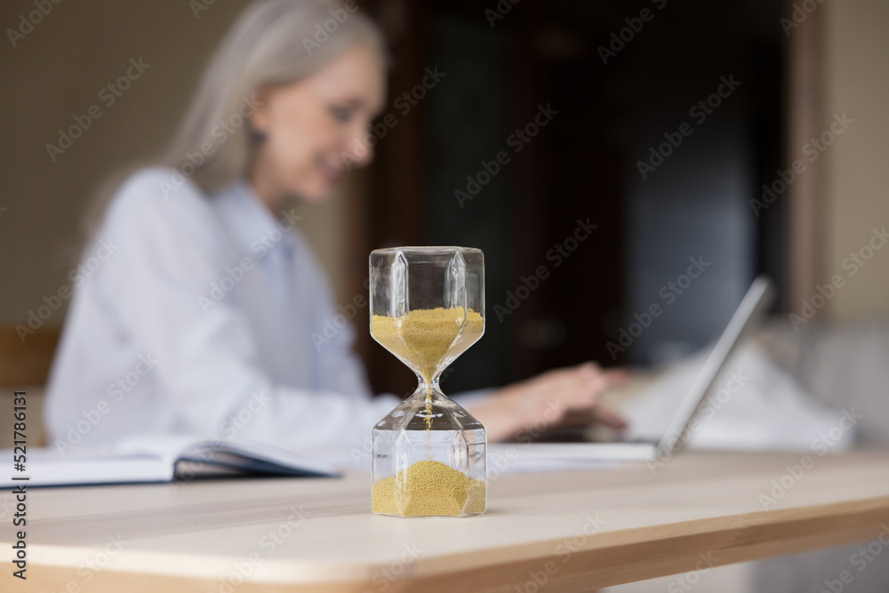 Hourglass with flowing sand on work desk of elderly businesswoman, closed up object. Mature business professional lady typing on laptop in blurred background. Career, time management concept