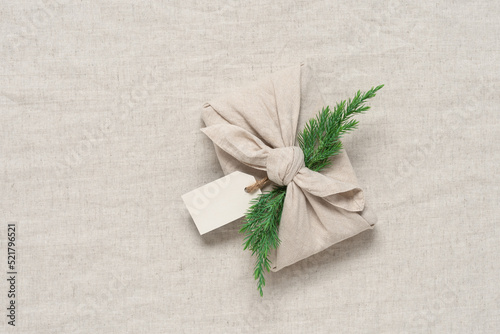 Christmas gift wrapped in fabric with juniper, beige linen textile background. A traditional Japanese furoshiki gift. Zero waste concept. Top view, selective focus, flat lay