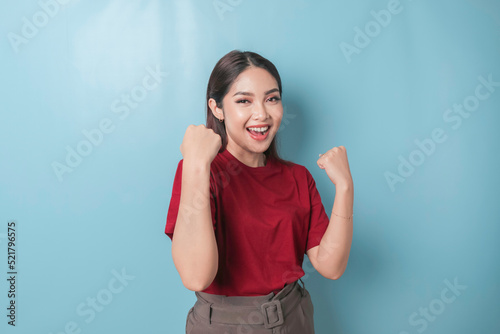 A young Asian woman with a happy successful expression wearing a red t-shirt isolated by blue background © Reezky