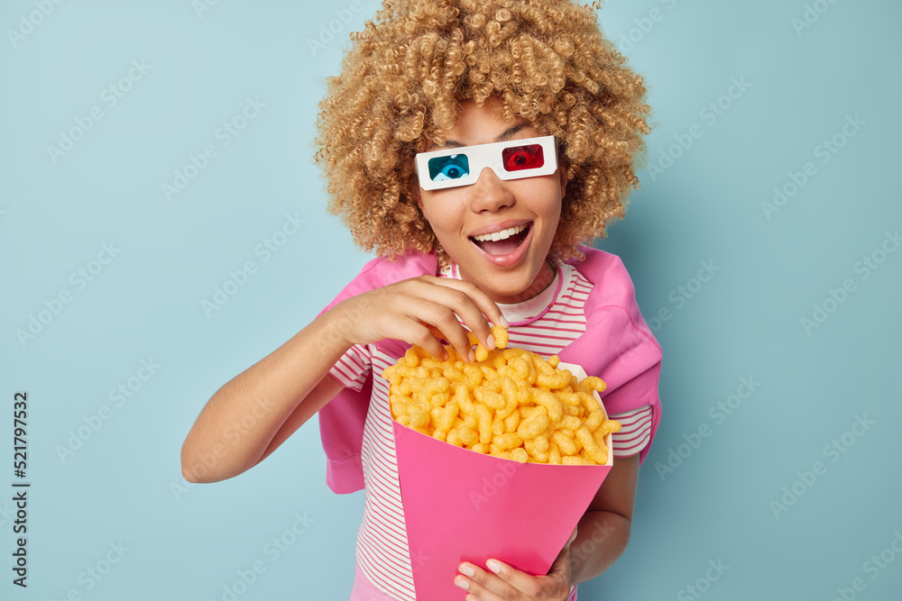 Obraz premium Surprised cheerful woman with curly bushy hair watches movie wears cinema eyeglasses eats corn sticks dressed in casual clothes isolated over blue background. Female model enjoys favorite snack