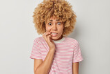 Studio shot of pretty young woman with curly hair bites finger nails looks anxious afraids of something wears earrings and striped t shirt suffers from panic attack isolated over grey background.