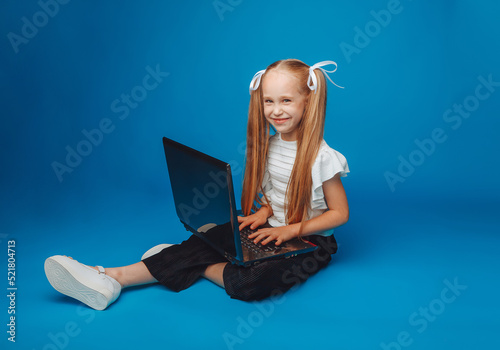 a schoolgirl is sitting on a blue background and looking at a laptop. a little girl is studying online. the child is typing on a laptop. isolated background.