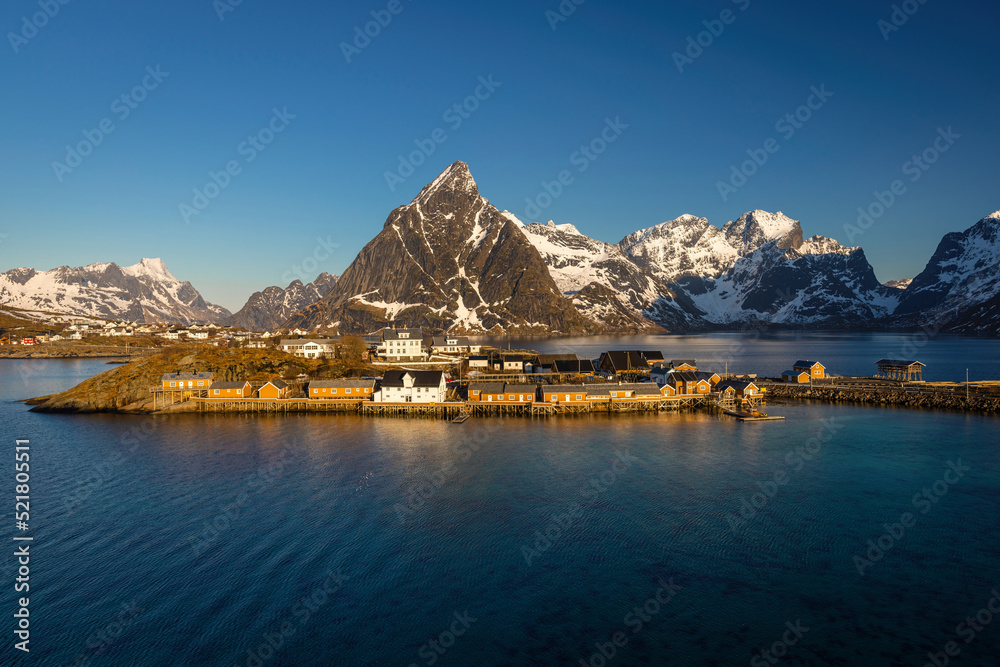 Norwegian fishing village with yellow buildings and snowy mountains on the background and sea on the foreground. Sunny weather with blue sky