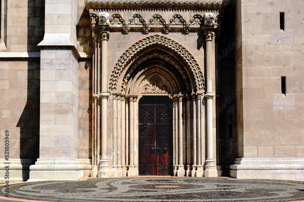 Naklejka premium Beautiful oak wood church main door or gate of the Matthias church in Budapest. black forged steel decorative leaf pattern. carved beige stone entrance in neo gothic style. rich ornate sculpted arch