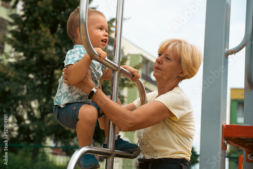 Grandmother supporting and holding her 2 years old grandson at children playground. Grandmother and child having fun. Support childhood symbol. Sunny, summer day