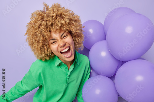 Overjoyed curly haired woman exclaims loudly feels very happy wears green shirt holds bunch of inflated balloons celebrates special occasion enjoys party time isolated over purple background