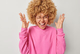 Positive woman with curly hair shouts loudly keeps palms raised reacts to awesome news wears spectacles and pink sweatshirt has overjoyed expression isolated over grey background. Human reactions