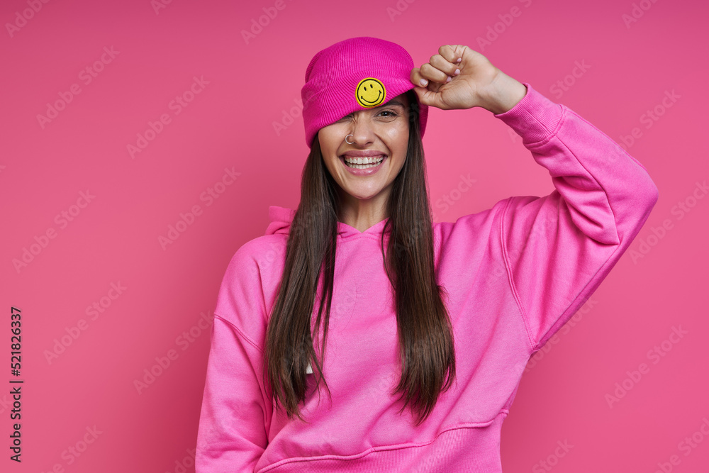 Happy young woman in hooded shirt adjusting her funky hat against pink background Happy young woman in hooded shirt adjusting her funky hat against pink background