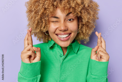 Beautiful curly haired woman hopes for better crosses fingers makes wish winks eye smiles broadly dressed in green shirt poses against purple background begs for dream come true. Body language