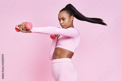 Confident young African woman exercising with dumbbells while standing against pink background © gstockstudio