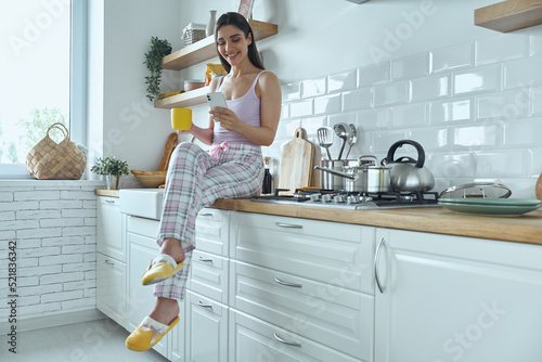 Relaxed young woman using smart phone and enjoying coffee while sitting on the kitchen counter