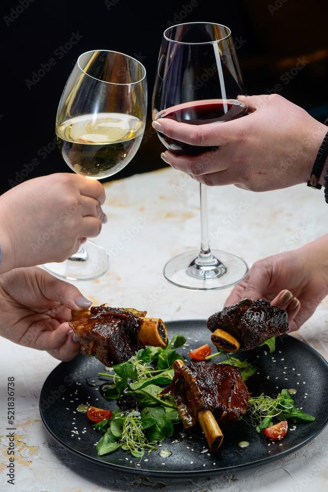 roast ribs in sauce in a black plate on a white background with wine glasses in hands vertical photo
